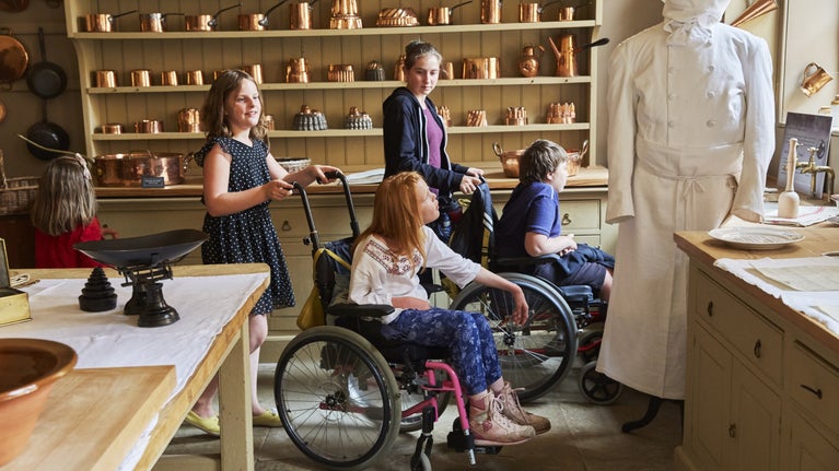 Two girls pushing two children (a girl and a boy) in wheelchairs in the kitchen at Attingham Park in Shropshire.